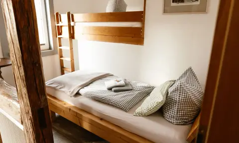 Einzelzimmer: Bett mit karierter Bettwäsche in der Holznische, darüber ein Hochbett